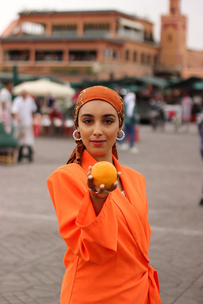 Mulher segurando uma laranja em uma praça movimentada, vestindo roupa laranja